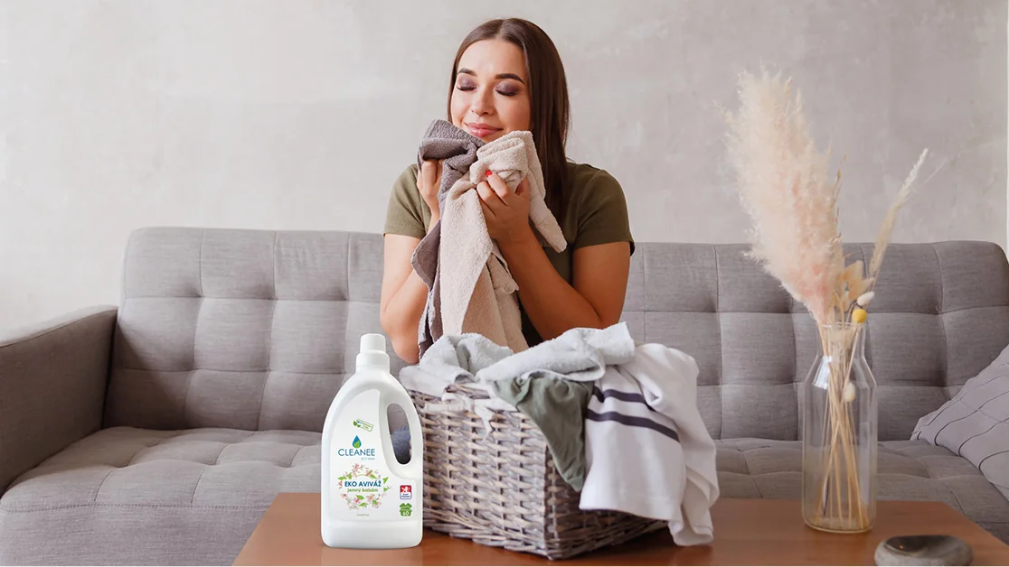 Žena si přivoní k čerstvě vypranému ručníku, vedle koš s prádlem a aviváž CLEANEE Jemný balzám 1,5 l